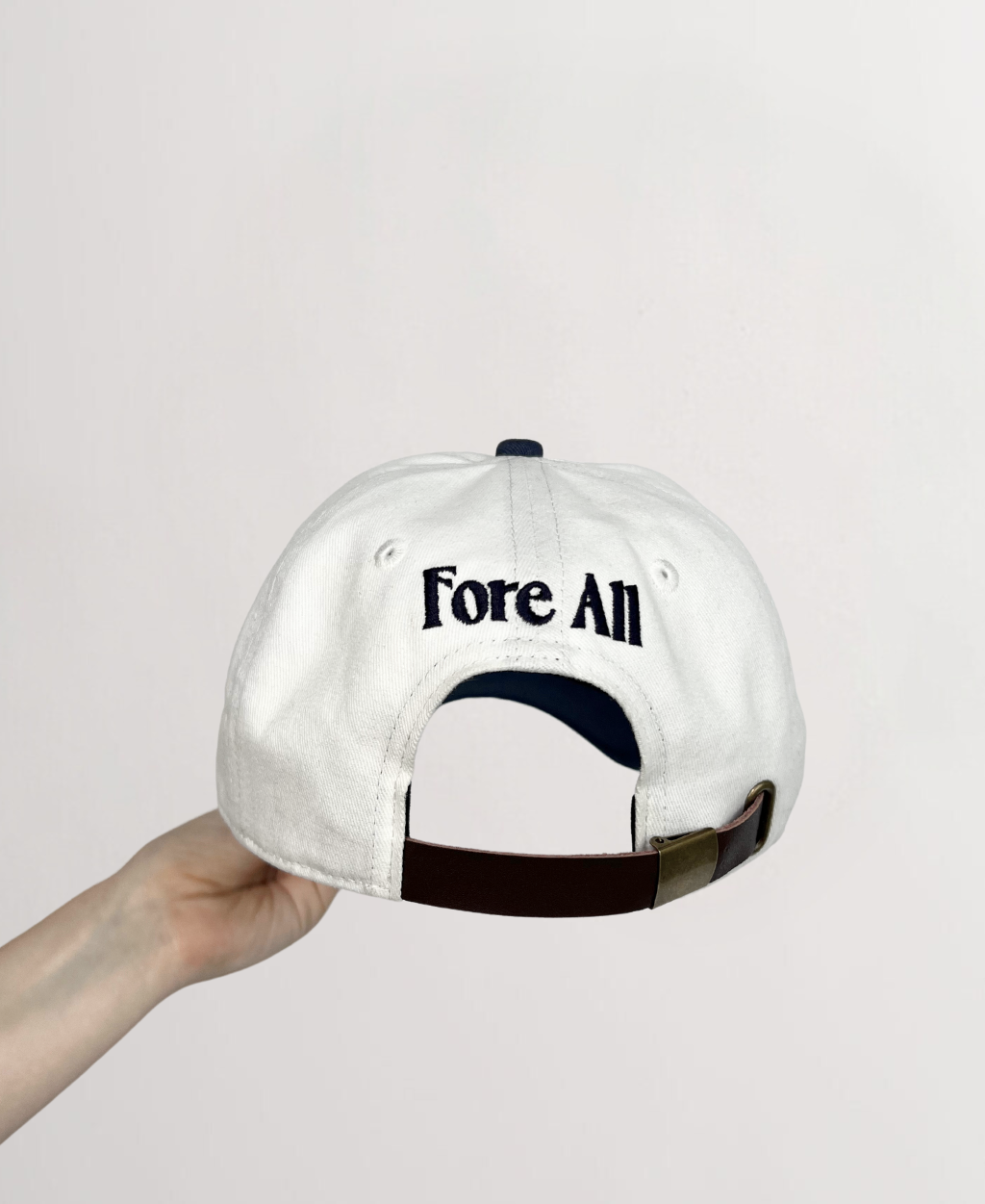 Back of white ball cap with indigo stitching reading &quot;Fore All&quot;  and faux leather strap attached to buckle for adjustments